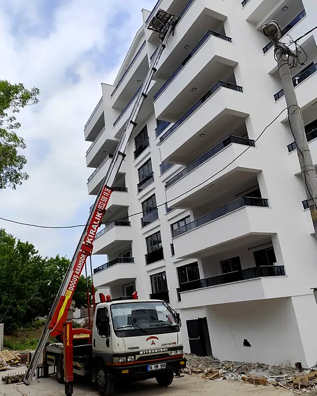 Yeni bir apartmanın önüne kurulmuş, kırmızı asansör kolu cepheye uzanan Doğuş asansörlü aracı